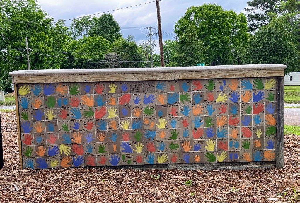 A colorful wall display featuring handprints in various colors, symbolizing community unity and participation, surrounded by greenery and utility poles.